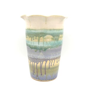 May include: A ceramic vase with a flared, wavy rim. The exterior features a layered glaze design with a cream-colored top section transitioning into horizontal bands of sage green, light blue, and lavender, with vertical drips running downward.
