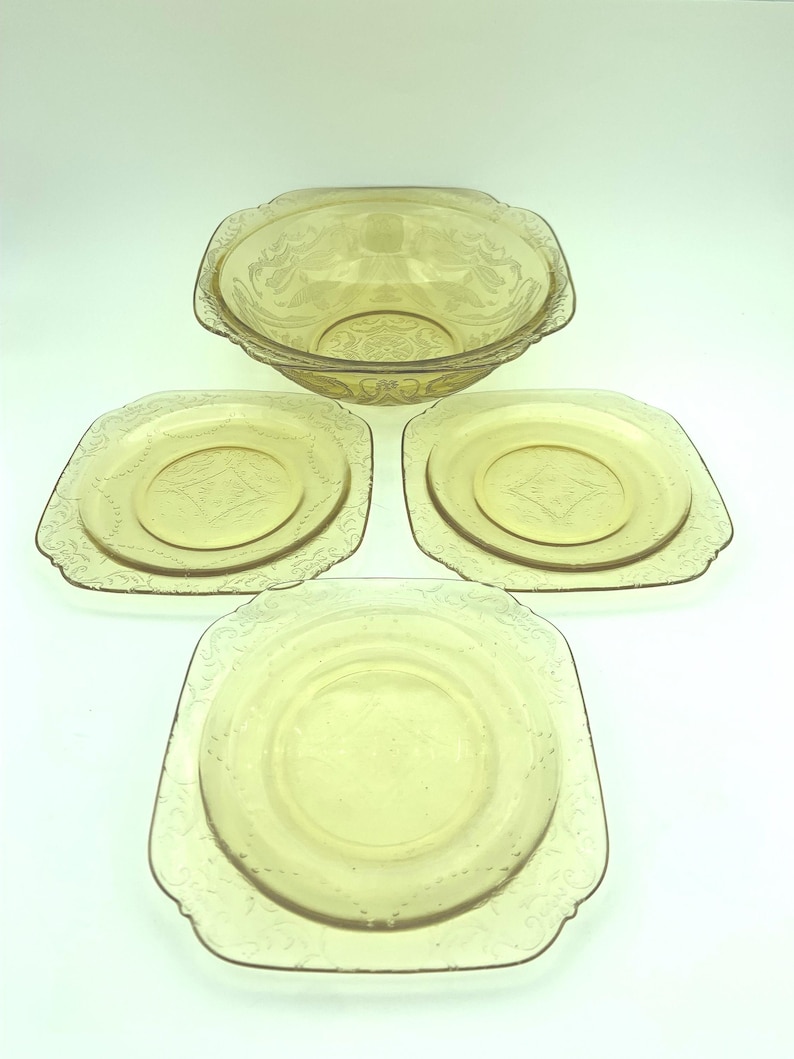 Puede incluir: Un juego de fuentes de vidrio color &aacute;mbar vintage. El juego incluye un cuenco grande y tres platos cuadrados. Cada pieza presenta un patr&oacute;n floral en relieve decorativo alrededor de los bordes y el centro. Los platos est&aacute;n colocados sobre una superficie blanca.