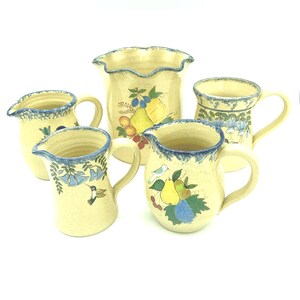 May include: A collection of five cream-colored ceramic vessels featuring hand-painted fruit, floral, and hummingbird motifs. Each piece has a speckled finish and a blue-toned rim. The set includes two pitchers, two mugs, and one vase.
