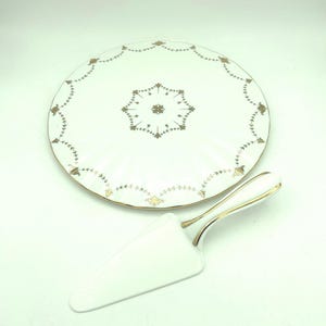 May include: A white porcelain cake plate and matching cake server. The plate features a scalloped edge with a gold-toned floral and vine pattern, and a central star-shaped gold motif. The server has a white triangular blade and a gold-trimmed handle.