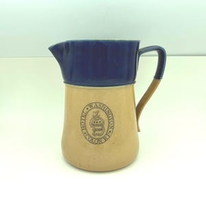 May include: A vintage ceramic pitcher with a tan base and a dark blue rim and handle. The pitcher features a circular emblem with the text "HOTEL WASHINGTON COLON RP". The handle has a tan accent. The pitcher is likely used for serving beverages.
