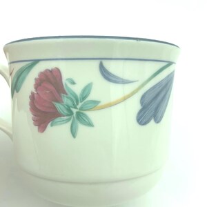 Puede incluir: Una taza de cer&aacute;mica de color crema con un patr&oacute;n floral pintado que incluye una flor de color rojo oscuro, hojas verdes y p&eacute;talos azules estilizados. El borde est&aacute; acentuado con una fina l&iacute;nea azul y el asa es parcialmente visible a la izquierda.