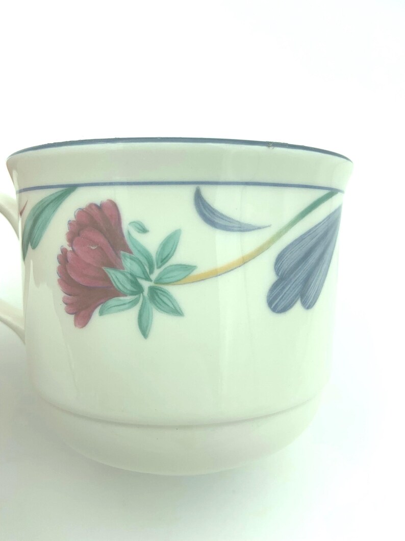 Puede incluir: Una taza de cer&aacute;mica de color crema con un patr&oacute;n floral pintado que incluye una flor de color rojo oscuro, hojas verdes y p&eacute;talos azules estilizados. El borde est&aacute; acentuado con una fina l&iacute;nea azul y el asa es parcialmente visible a la izquierda.