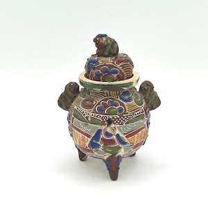 May include: A decorative, multi-colored ceramic incense burner with a lid and three legs. The burner features intricate painted designs, including figures and floral patterns, with a small animal figure on top.