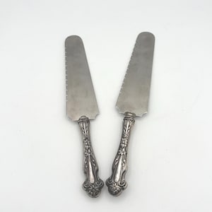May include: Two silver cake knives with serrated blades and ornate floral handles. The knives are displayed against a white background, showcasing their elegant design. Ideal for serving cake at special events.