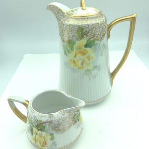 May include: A white porcelain tea set featuring a tall teapot with a lid and a small creamer. The set is decorated with yellow roses, gold trim, and a textured, dotted pattern. The teapot has a gold handle and spout.