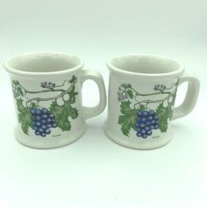 May include: Two white ceramic mugs with a handle. Each mug features a blue grape cluster design with green leaves and the word "Vine". The mugs are on a white background.