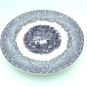 May include: A round, antique plate with a detailed blue and white landscape scene in the center. The plate has a decorative border with floral and scroll patterns. The scene depicts trees, buildings, and swans on a body of water.