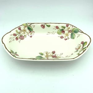 May include: An off-white ceramic serving dish with an octagonal shape and a brown rim. The interior features a botanical pattern of white apple blossoms, green leaves, and small reddish-brown buds arranged along the edges of the shallow bowl.