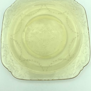 Puede incluir: Un plato cuadrado de vidrio amarillo p&aacute;lido con un borde decorativo y patrones florales y geom&eacute;tricos grabados. El plato tiene un centro ligeramente elevado y una est&eacute;tica vintage delicada.