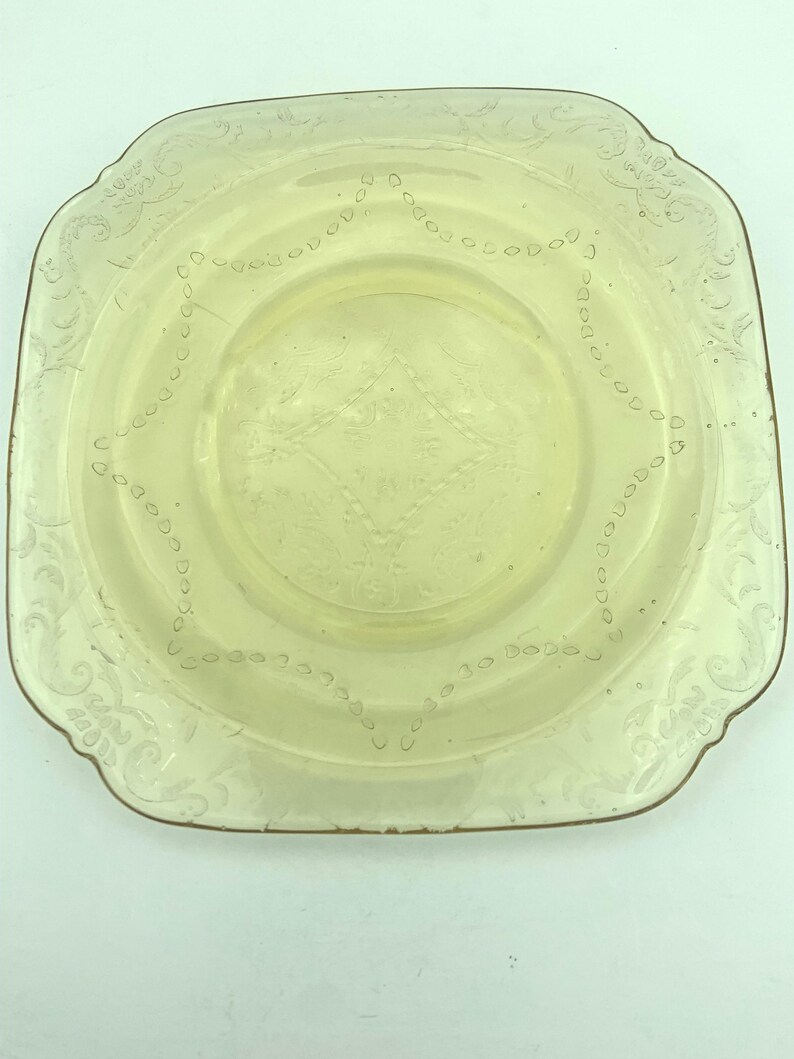 Puede incluir: Un plato cuadrado de vidrio amarillo p&aacute;lido con un borde decorativo y patrones florales y geom&eacute;tricos grabados. El plato tiene un centro ligeramente elevado y una est&eacute;tica vintage delicada.