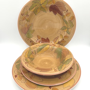 May include: A set of four ceramic plates and bowls. The plates and bowls are tan with a leaf design in shades of green, brown, and yellow. The rims are a reddish-brown color. The set includes two dinner plates and two smaller bowls.