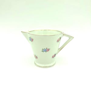 May include: A white porcelain creamer featuring a delicate floral pattern of small pink and blue roses with green leaves scattered across the exterior. The vessel has a gold-trimmed rim, a triangular spout, and an angular handle on the side.