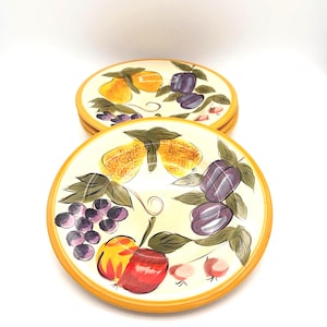 May include: A stack of round ceramic plates featuring a hand-painted fruit motif on a cream background. The design includes yellow pears, purple plums, a cluster of dark grapes, and a red apple, all surrounded by green leaves and a yellow rim.