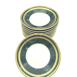 May include: A stack of round plates featuring a white center, a dark green marble-patterned border, and a gold-colored rim with a decorative geometric pattern. The plates are arranged vertically, showing the top plate clearly in the foreground.