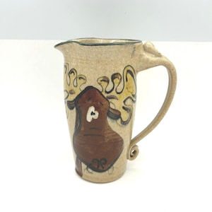 May include: Tan ceramic pitcher with a brown and yellow abstract design. The pitcher has a curved handle and a spout for pouring. The design features a brown shape with a white accent and yellow swirls.