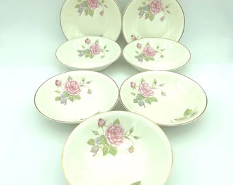 May include: A set of seven cream-colored ceramic bowls featuring a delicate pink rose and green leaf floral pattern in the center. Each bowl has a smooth, rounded edge with a thin gold-tone trim along the rim, creating a uniform appearance.