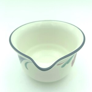 Puede incluir: Un cuenco vertedor de cer&aacute;mica de color crema con un borde azul oscuro y un pico distintivo. La superficie exterior est&aacute; decorada con un sutil patr&oacute;n floral en tonos apagados de verde y rojo, ofreciendo un recipiente de cocina funcional.