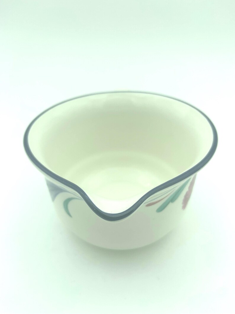 Puede incluir: Un cuenco vertedor de cer&aacute;mica de color crema con un borde azul oscuro y un pico distintivo. La superficie exterior est&aacute; decorada con un sutil patr&oacute;n floral en tonos apagados de verde y rojo, ofreciendo un recipiente de cocina funcional.