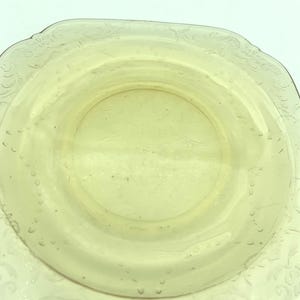 Puede incluir: Un plato de vidrio amarillo p&aacute;lido, de forma cuadrada, con un centro ligeramente elevado. El plato tiene un delicado patr&oacute;n floral en relieve alrededor del borde y peque&ntilde;os detalles en forma de burbujas. El dise&ntilde;o general es simple y elegante.