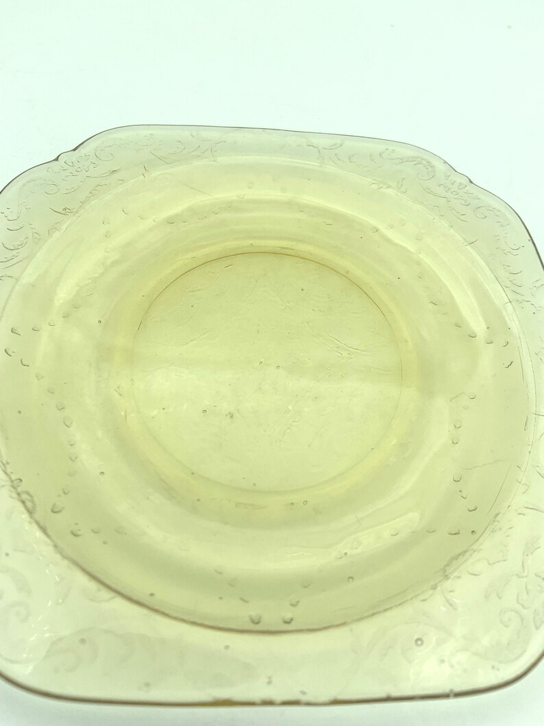 Puede incluir: Un plato de vidrio amarillo p&aacute;lido, de forma cuadrada, con un centro ligeramente elevado. El plato tiene un delicado patr&oacute;n floral en relieve alrededor del borde y peque&ntilde;os detalles en forma de burbujas. El dise&ntilde;o general es simple y elegante.