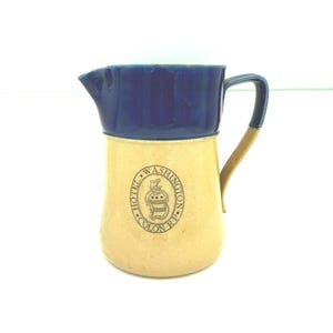 May include: A two-toned ceramic pitcher featuring a dark blue upper section and a tan lower body. The front displays a circular logo with the text HOTEL WASHINGTON R.P. and a central crest emblem. It includes a curved handle and a pouring spout.