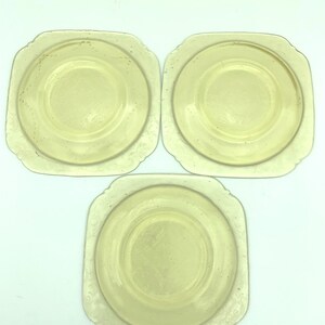 Puede incluir: Tres platos antiguos de vidrio amarillo. Cada plato tiene forma cuadrada con esquinas redondeadas y un centro circular. Los bordes presentan un delicado dise&ntilde;o floral en relieve. Los platos est&aacute;n dispuestos sobre un fondo blanco.