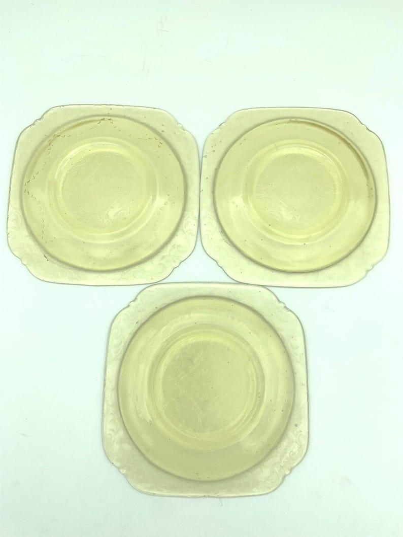 Puede incluir: Tres platos antiguos de vidrio amarillo. Cada plato tiene forma cuadrada con esquinas redondeadas y un centro circular. Los bordes presentan un delicado dise&ntilde;o floral en relieve. Los platos est&aacute;n dispuestos sobre un fondo blanco.