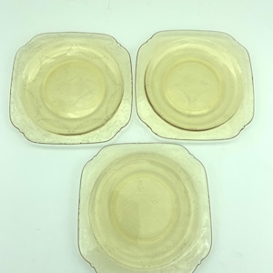 Puede incluir: Tres platos vintage de vidrio amarillo. Cada plato tiene forma cuadrada con esquinas redondeadas, un patr&oacute;n central de diamante y un borde decorativo. Los platos est&aacute;n dispuestos sobre una superficie blanca.