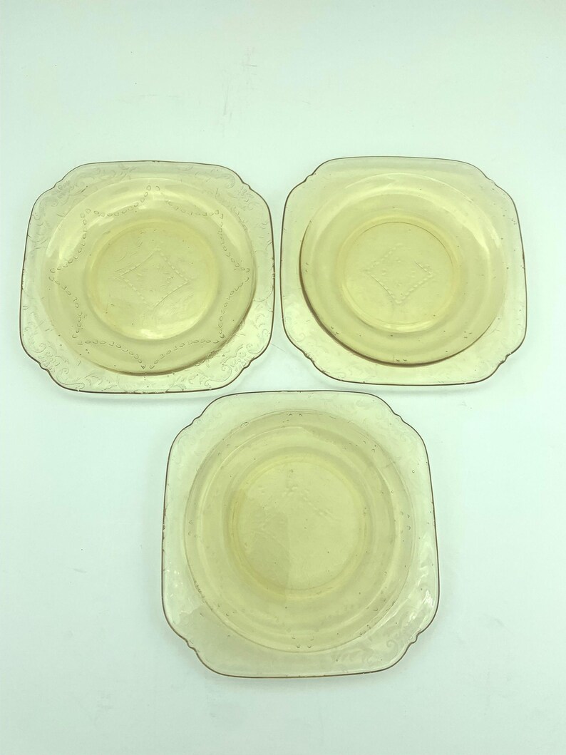 Puede incluir: Tres platos vintage de vidrio amarillo. Cada plato tiene forma cuadrada con esquinas redondeadas, un patr&oacute;n central de diamante y un borde decorativo. Los platos est&aacute;n dispuestos sobre una superficie blanca.