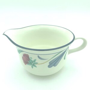 Puede incluir: Una jarrita de cer&aacute;mica color crema con un borde azul oscuro y un patr&oacute;n floral que incluye una rosa roja y hojas azules estilizadas. La jarrita tiene un asa curva en el lado derecho y un pico vertedor en el lado izquierdo del recipiente.