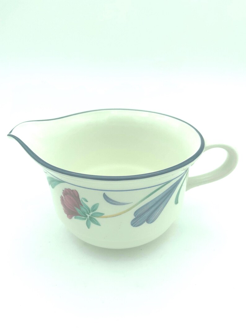 Puede incluir: Una jarrita de cer&aacute;mica color crema con un borde azul oscuro y un patr&oacute;n floral que incluye una rosa roja y hojas azules estilizadas. La jarrita tiene un asa curva en el lado derecho y un pico vertedor en el lado izquierdo del recipiente.