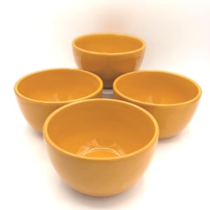 May include: A set of four matching mustard yellow bowls with a smooth, glossy finish. Each bowl features a rounded shape with a wide opening and a deep interior, arranged in a cluster to display their uniform color and consistent design.