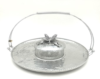 May include: A round, hammered silver-tone metal serving tray with a floral embossed design and a tall, arched wire handle. Centered on the tray is a small, round glass dish with a silver-tone metal lid topped with a sculpted metal flower.