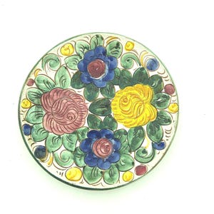 May include: A round ceramic plate featuring a hand-painted floral design. The pattern includes two blue flowers, one pink rose, and one yellow rose, all surrounded by green leaves and small yellow and blue dots on a white background.