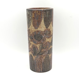 May include: A tall, cylindrical ceramic vase with a textured surface. The vase features a floral design with brown and green accents on a beige background. The vase has a narrow opening and a dark brown rim.