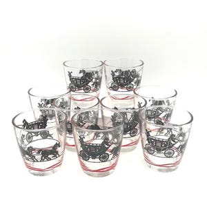 May include: A set of nine clear glass shot glasses, each featuring a black graphic illustration of a horse-drawn carriage and a red wavy line accent near the base. The glasses are arranged in a group, showcasing the repeating carriage design.