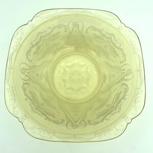 Puede incluir: Un cuenco de vidrio amarillo p&aacute;lido con forma cuadrada y bordes festoneados. El cuenco presenta un dise&ntilde;o floral y geom&eacute;trico grabado. El centro del cuenco tiene un patr&oacute;n circular. El cuenco es probablemente vintage o antiguo.