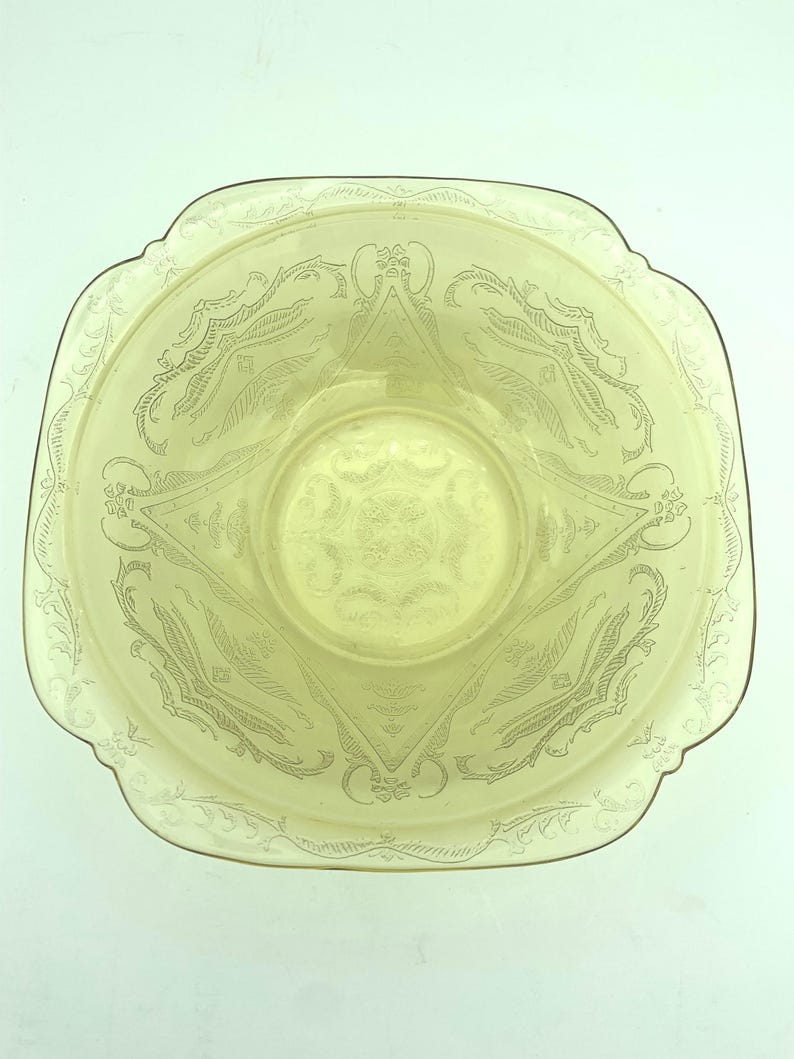 Puede incluir: Un cuenco de vidrio amarillo p&aacute;lido con forma cuadrada y bordes festoneados. El cuenco presenta un dise&ntilde;o floral y geom&eacute;trico grabado. El centro del cuenco tiene un patr&oacute;n circular. El cuenco es probablemente vintage o antiguo.