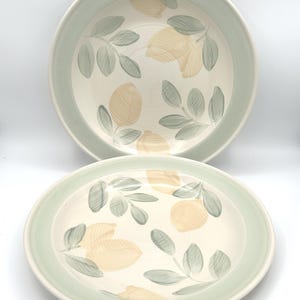 May include: Two ceramic plates with a cream-colored interior and a light green rim. The plates feature a hand-painted design of lemons and green leaves. The lemons are a soft yellow color, and the leaves are a muted green.