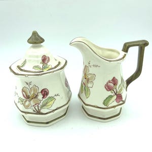 May include: A cream-colored ceramic sugar bowl with a matching lid and a creamer pitcher. Both pieces feature a hand-painted floral pattern with pink and white blossoms, green leaves, and brown trim along the edges and the creamer handle.
