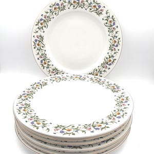 May include: A stack of white ceramic dinner plates featuring a delicate floral vine pattern along the rim. The pattern consists of green stems with small blue, yellow, and pink flowers. One plate stands upright behind the stack of five plates.