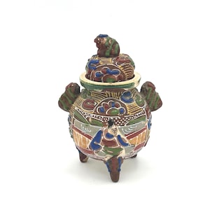 May include: A small, three-legged ceramic jar with a removable lid featuring a sculpted animal finial. The vessel is decorated with hand-painted figures in blue, red, green, and gold, set against a textured brown background with intricate patterns.