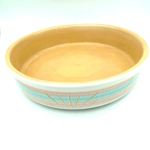 May include: An oval ceramic baking dish with a smooth, light tan interior. The exterior features horizontal bands of soft pink and teal glaze, accented by a carved sunburst pattern that reveals the white clay body beneath the colorful glaze.