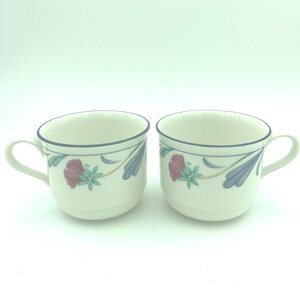 Puede incluir: Dos tazas de cer&aacute;mica color blanco hueso con un delicado patr&oacute;n floral de p&eacute;talos rojos y azules, hojas verdes y l&iacute;neas azules finas. Cada taza tiene un asa curva y un cuerpo redondeado, mostrando un dise&ntilde;o consistente en ambas.