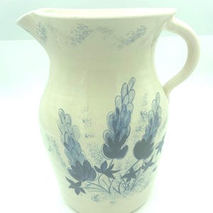 May include: A cream-colored ceramic pitcher featuring a hand-painted blue floral pattern of lupine-like flowers. The pitcher has a curved handle, a wide spout, and a speckled blue design around the rim and base of the floral motif.