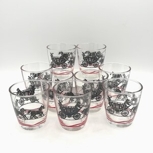 May include: Set of nine clear glass tumblers. Each tumbler features a black illustration of a horse-drawn carriage with a red stripe near the base. The glasses are arranged in a group on a white surface.