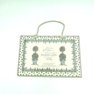May include: A rectangular ceramic wall plaque with a rope hanger, featuring a cream background, a border of green leaves and red berries, and two potted topiary illustrations flanking the text: Each day is God's Gift to us. We must make it blossom and grow into a thing of beauty.