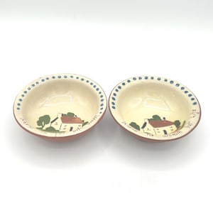 May include: Two small, round ceramic bowls with a cream-colored interior and a reddish-brown exterior. Each bowl features a hand-painted scene of a house and trees, with blue dots around the rim. Text is visible on the interior of the bowls.