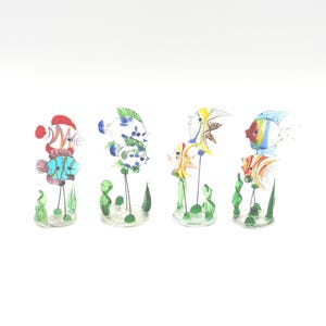 May include: Four colorful glass fish sculptures with detailed patterns and designs. Each fish is mounted on a clear base with green glass seaweed accents. The fish feature various colors, including red, blue, yellow, and orange.
